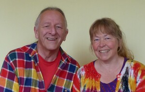 A smiling older couple posing for a photo indoors against a pale yellow wall, wearing colorful shirts.
